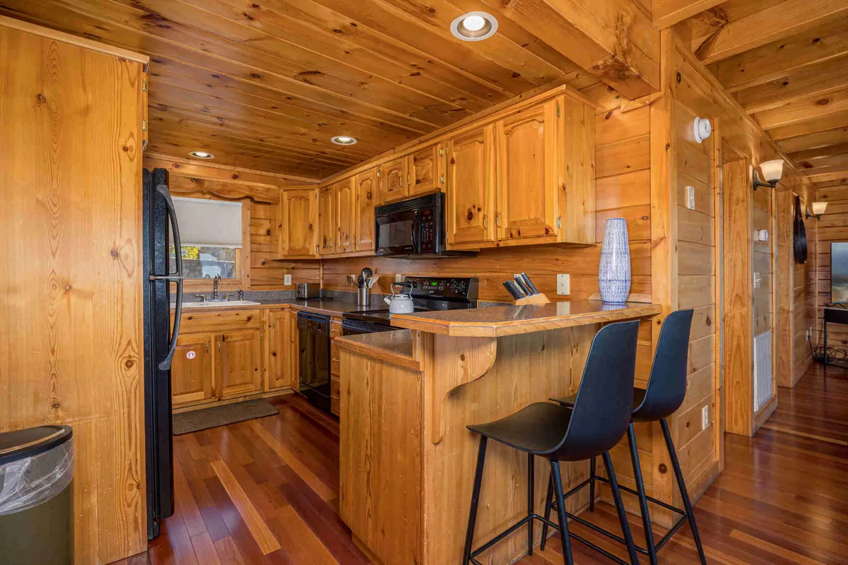 This image showcases a modern kitchen featuring wooden cabinetry, sleek black appliances, and a spacious countertop with bar stools. The space exudes warmth and functionality, inviting both cooking and socializing. The open cabinetry and bar seating suggest an inviting atmosphere for meal preparation and conversation.