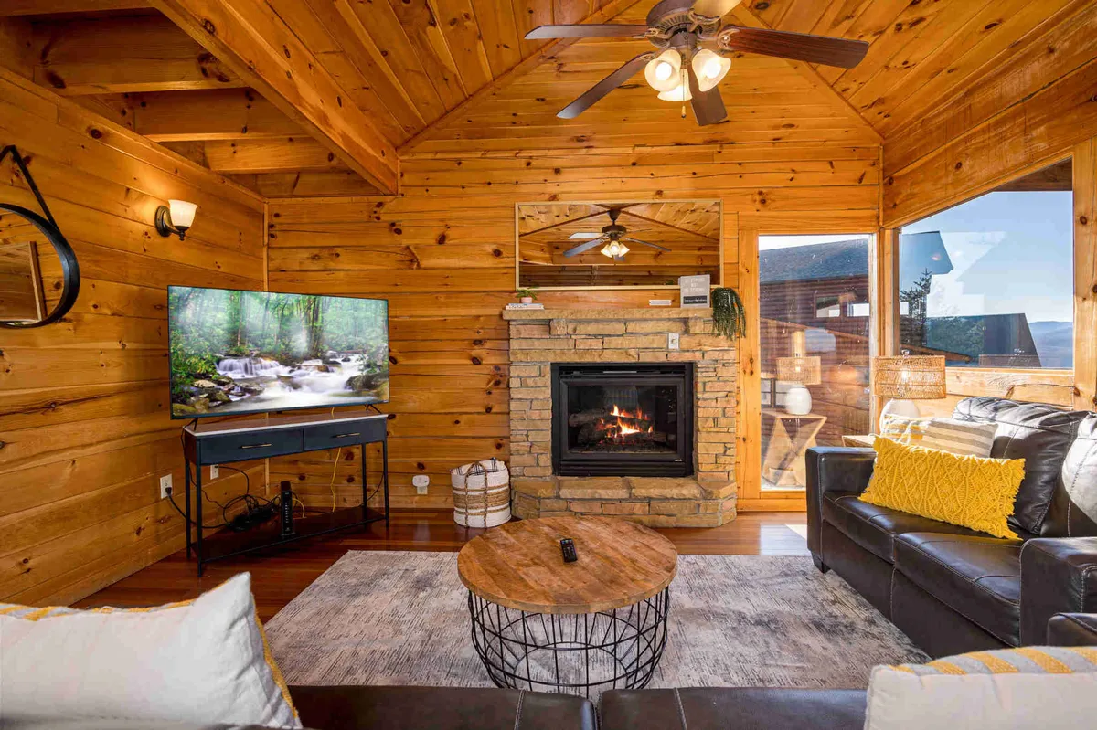 A warm living room setting featuring a stone fireplace, wooden ceiling beams, a large TV, a coffee table, and a comfortable leather couch. The rustic woodwork and plush couches evoke a cozy, inviting atmosphere ideal for gatherings.