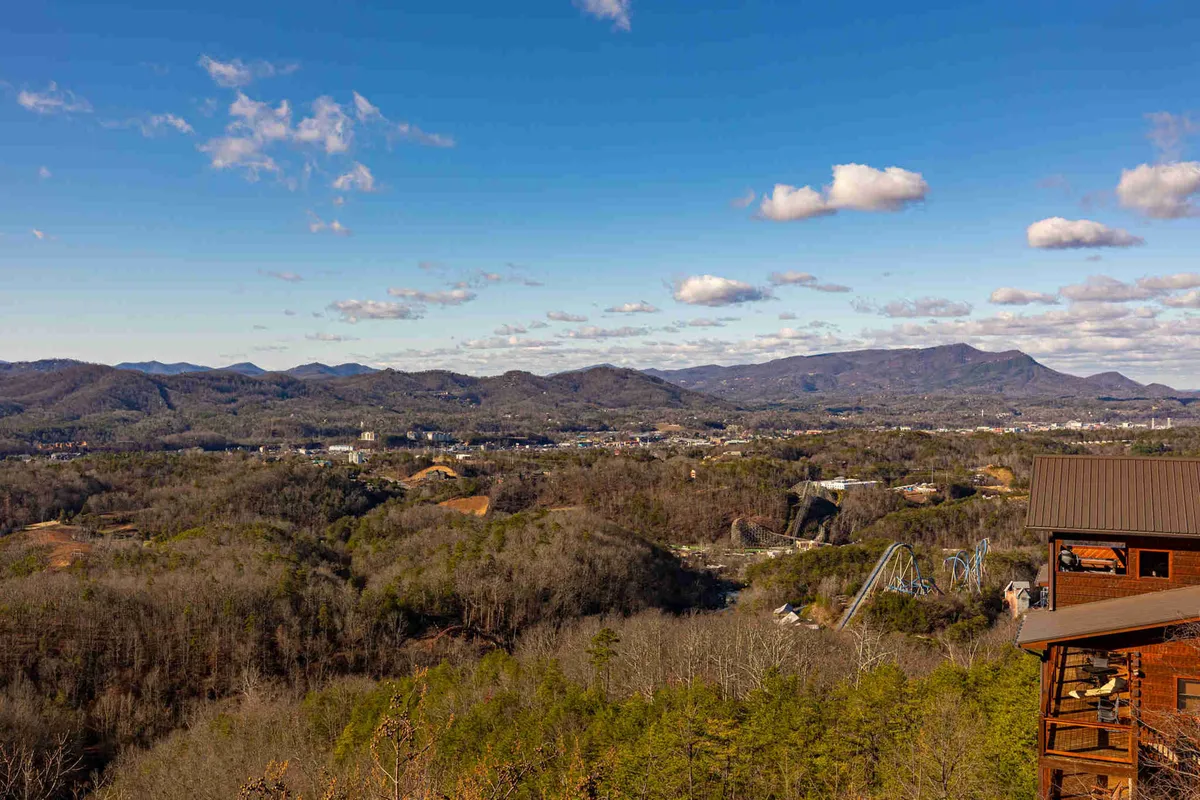 A wide, sweeping view of the Smoky Mountains, with layers of mountain range stretching into the distance, dense forests below, and a hint of cityscape and a roller coaster for local flavor. The natural light and open sky invite viewers to imagine hiking, sightseeing, or indulging in nature photography.