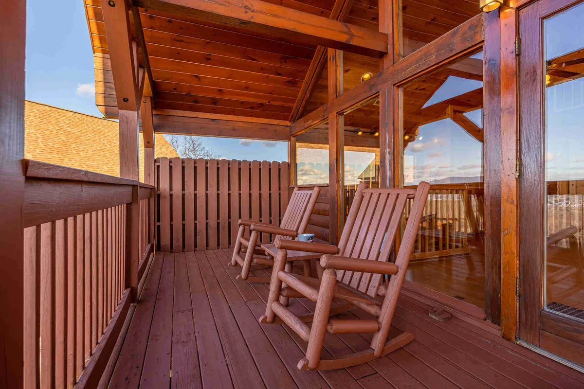 A serene exterior image of a wooden deck furnished with rocking chairs, large glass doors opening to the cabin, and expansive views of the surrounding mountains beyond. The inviting deck space implies relaxation after a day spent outdoors.