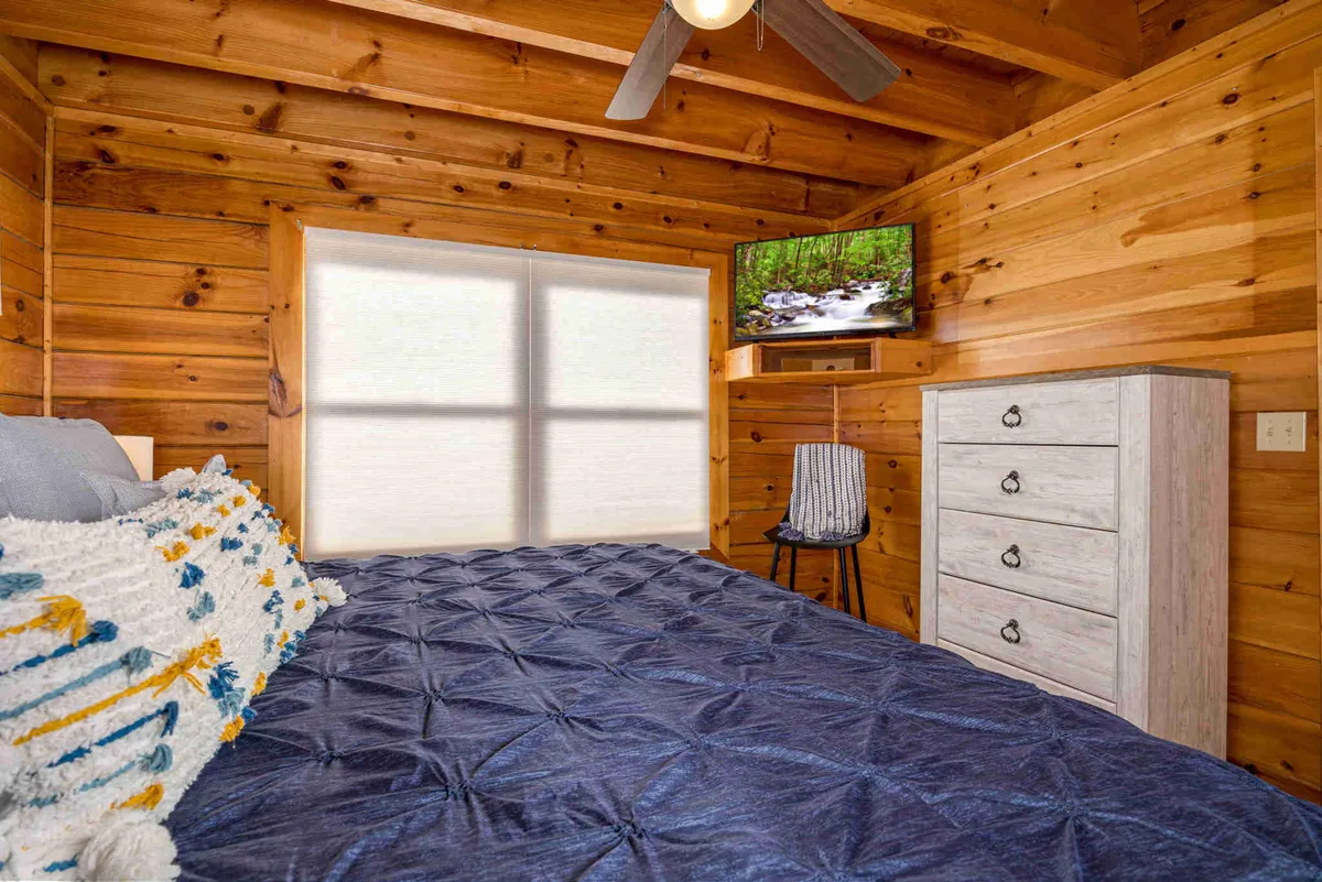 A bedroom with a textured blanket on the bed, warm wooden walls, a dresser, and a TV displaying a calming nature scene. Natural light streams into the space, reinforcing a sense of serenity and comfort.