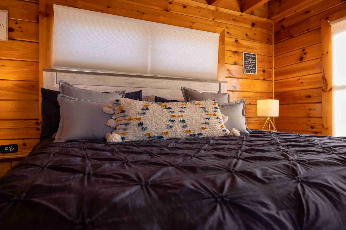 A cozy bedroom featuring plush bedding with decorative pillows, wooden walls, and a bedside lamp. Soft natural light filters into the room, highlighting the tranquil, inviting atmosphere ideal for relaxation and restful sleep.
