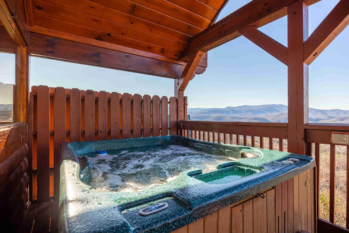 A private hot tub set on a wooden deck with an expansive view of the Smoky Mountains in the background. The setting exudes tranquility, with the hot tub oriented to maximize the sweeping mountain vistas. A portion of the wooden cabin’s structure frames the scene, suggesting privacy and rustic comfort.