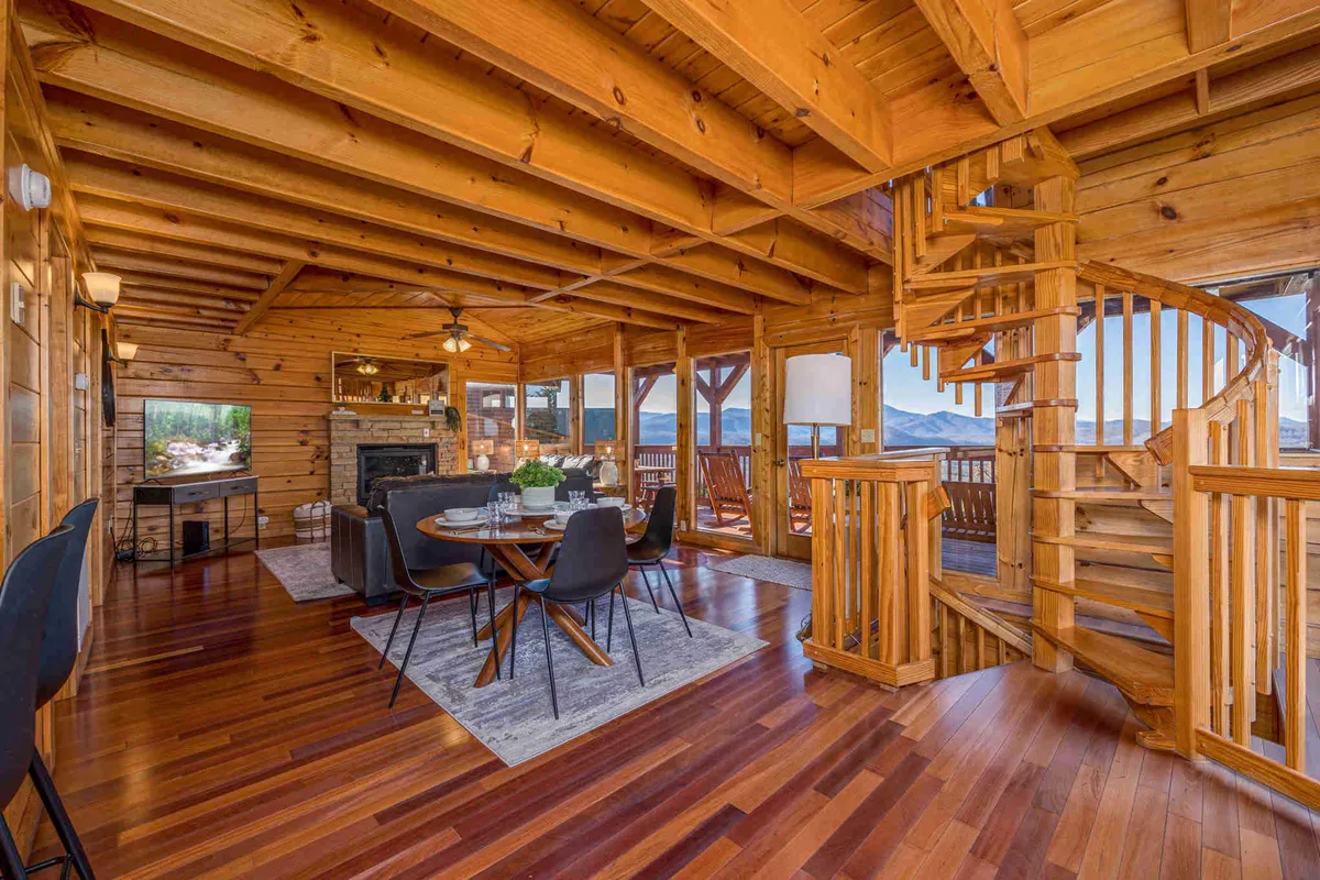 An open dining space features a wooden spiral staircase, a dining table set for guests, a cozy stone fireplace, and expansive windows offering breathtaking mountain views. Natural light fills the room, accentuating the welcoming, open-concept layout.