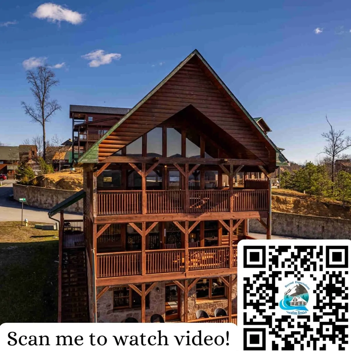 The image showcases a stunning, large wooden cabin with multiple balconies, set against a backdrop of clear blue skies. Expansive decks offer plenty of space for outdoor relaxation and family gatherings, inviting guests to enjoy sunrise views or stargaze in the evening. The setting suggests a tranquil, elevated perch overlooking the Smoky Mountains.